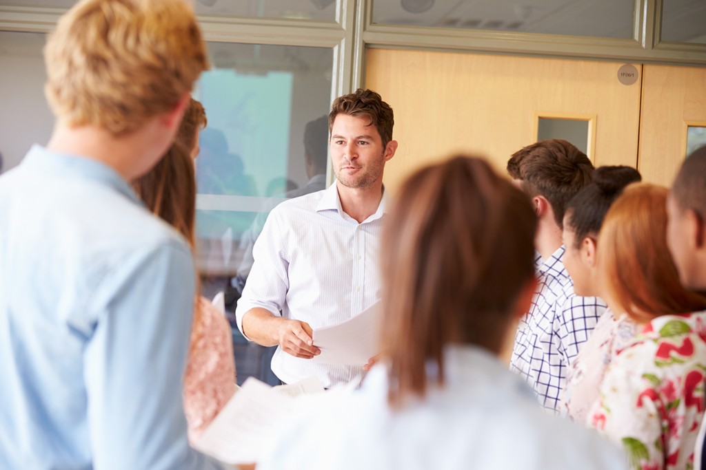 Teacher With College Students Standing By Desks In Classroom